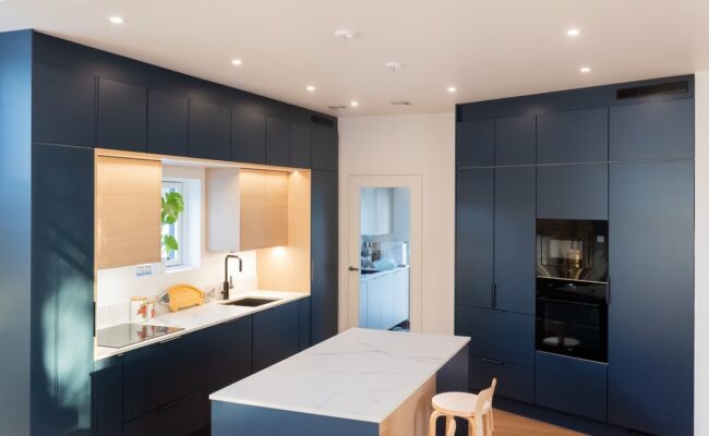 Tyylikäs keittiö, jossa on tummansiniset kaapistot ja valkoinen marmoritaso. Tilassa on ikkuna, jonka vieressä on viherkasvi ruukussa, sekä induktioliesi, uuni ja pieni puinen jakkara. Upotettu LED-valaistus valaisee tilan kirkkaasti. Tämä kaikki löytyy LedStore.fi:n laajasta LED-valaistusvalikoimasta!