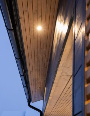 Viistostatu moderni talo, jossa tyylikäs puupaneelikatto, upotetut LED-pihavalot ja musta räystäskokuvat iltahämärässä kuvaruu LED-valaistuksen näyttävää ja nykyaikaista ilmettä. - LedStore LED spotlights in the eaves are installed to illuminate the exterior wall.