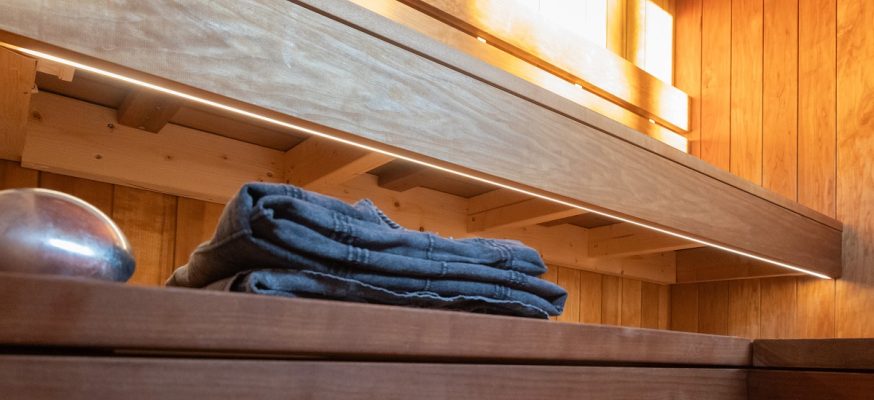 GZ9A7712 - LedStore Kodikkaan saunan puupinnan tunnelmaa täydentää pieni ikkuna, josta tulvii auringonvaloa. LedStore.fi:n himmeä LED-valo sulautuu kauniisti luonnonvaloon, tehden rauhallisesta ilmapiiristä entistä miellyttävämmän. Seinustalla kulkee penkki, ja tummaksi taiteltu pyyhe on asetettu kiiltävän teräspallon viereen puiselle tasolle.