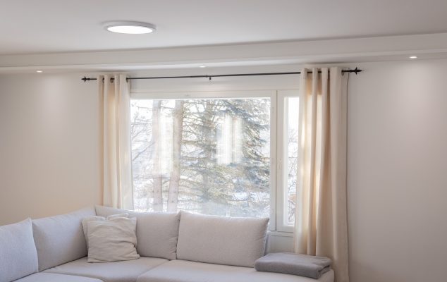 Infällda spotlights runt väggarna i vardagsrummet_LedStorefi - LedStore Minimalistinen olohuone LedStore.fi-tyyliin: Huoneessa on valkoinen kulmasohva, jota koristavat muutamat tyynyt. Suurten ikkunoiden kermanväriset verhot kehystävät kauniisti näkymää puustoon. Tunnelmallisen valaistuksen tarjoaa pehmeästi loistavat LED-kattovalot och upotetut LED-valaisimet, jotka luovat rauhallisen ilmapiirin koko tilaan.