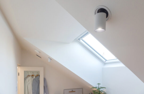 Minimalistinen huone vinolla katolla on valaistu pyöreällä LED-kattovalaisimella sekä kattoikkunalla, valikoimaa löytyy LedStore.fi-verkkokaupasta. Kattoikkunan alla sijaitsee pieni kasvi. Vasemmalla seinällä on osittain näkyvissä ovi, josta roikkuu vaatteita. Valkoiset seinät korostavat huomioita sisustusta ja luovatin ilmapiirin.