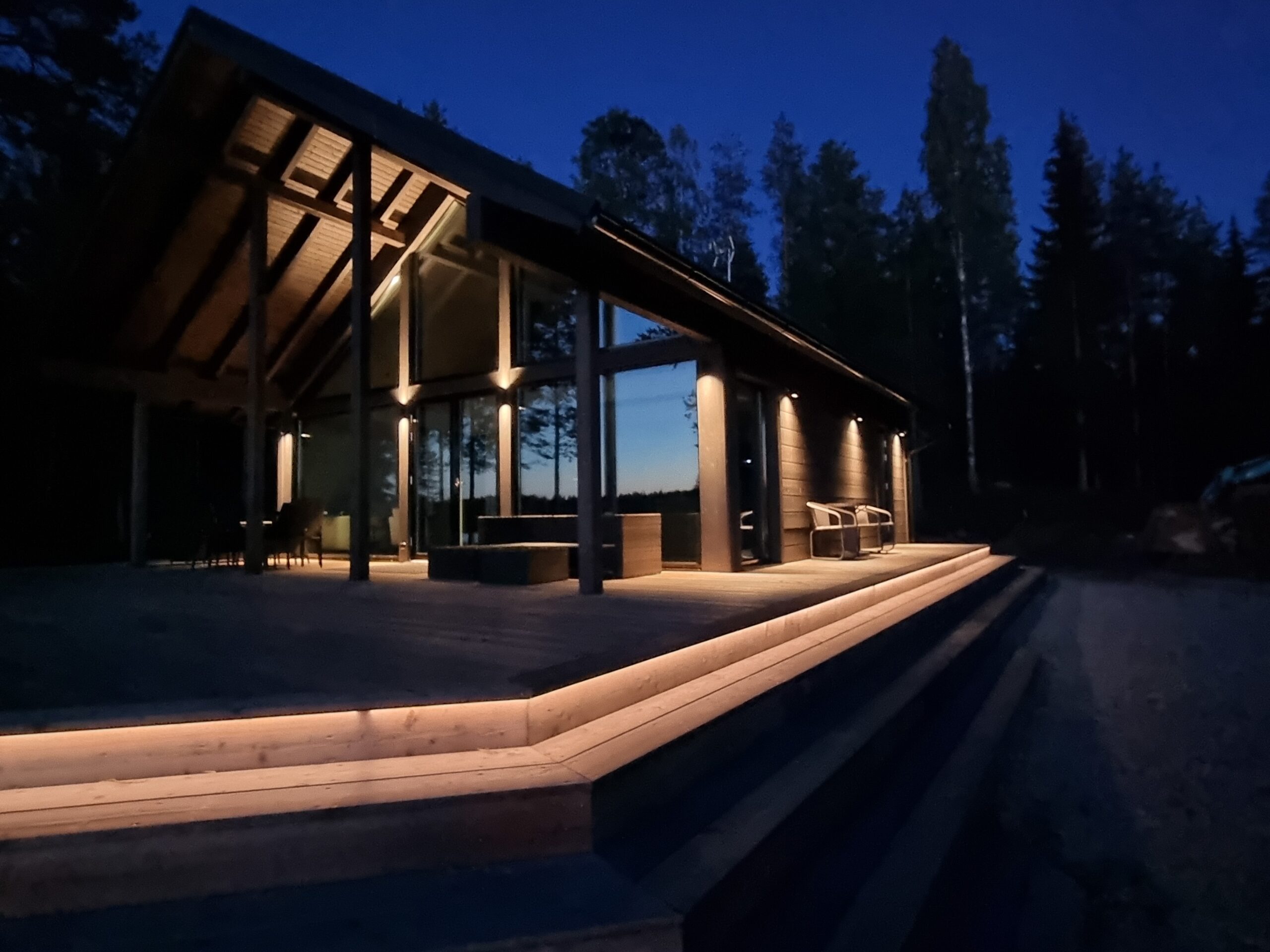 Nykyaikainen hirsimökki, jossa on suuret ikkunat ja ulkoportaat, kylpee lämpimässä tunnelmavalossa LedStore.fi:n laadukkaan ulkovalaistuksen ja led-valonauhojen mukana. Tyylikkäät led-valaisimet korostavat mökin arkkitehtuuria sekä ympäriä luontoa, luoden kutsuvan ilmeen iltahämärässä. - LedStore Led-valonauhat voidaan asentaa pitkiäkin matkoja yhtenäisenä. Tässä on valaistu terassin kiertävät porrasaskelmat niin, että ensimmäisen askelman alle on laitettu led-nauhaa, niin valon tuoma kontrasti osoittaa kulkijalle, missä kohtaa ensimmäinen askelma on.