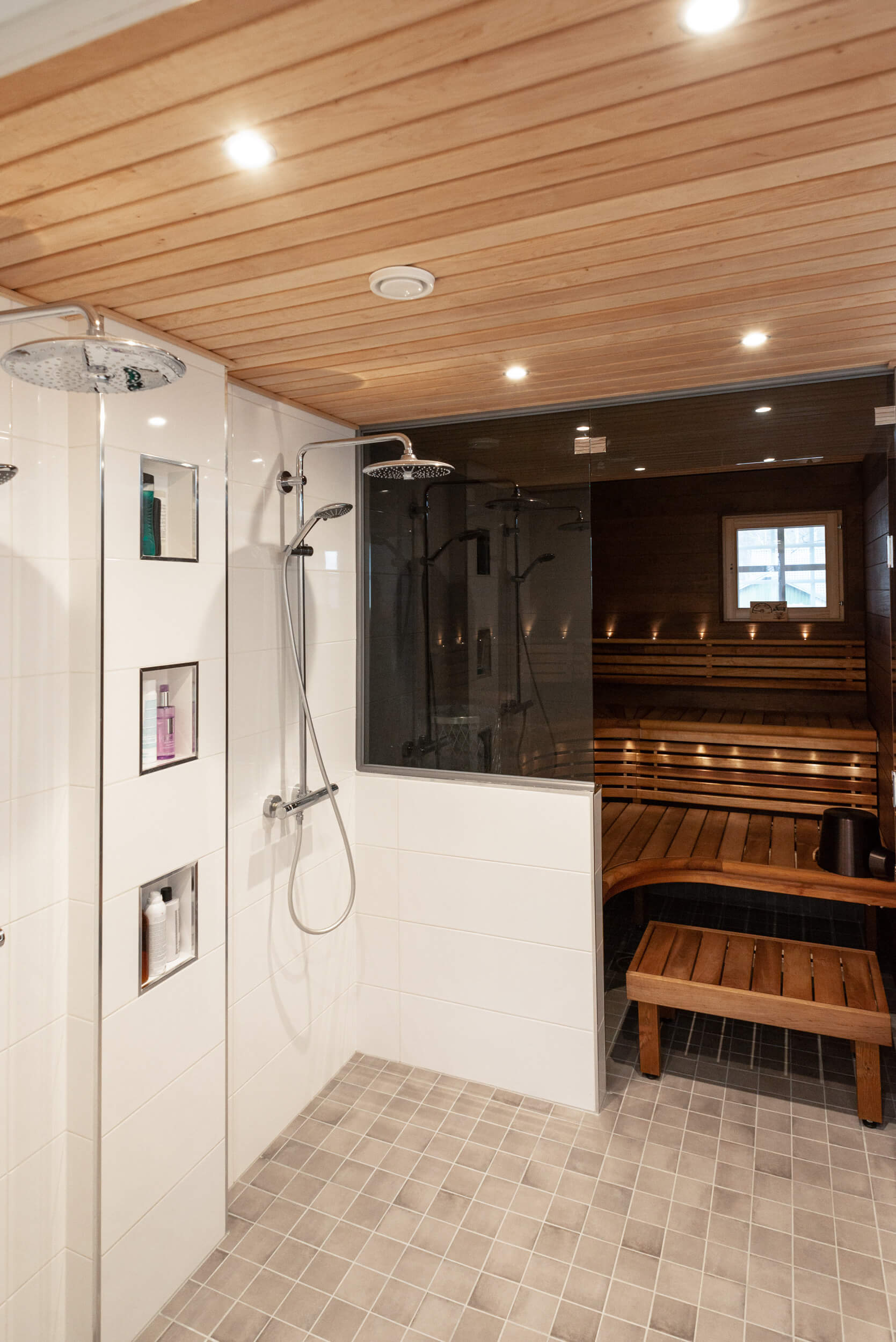 _DSC77265 - LedStore Moderni puusauna, jossa LED-valaistu kaakeloitu suihkutila, kaksi suihkupäätä, upotetut hyllyt ja lasiseinäiset lauteet pienen ikkunan vieressä.