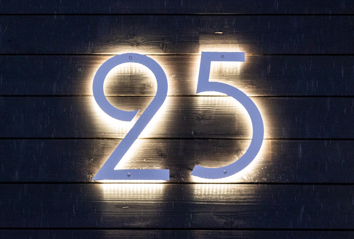 Valkoinen numero 25 tummalla puuseinällä, taustalta pehmeästi LedStore.fi:n LED-valaistuksella valaistuna, tuon ja modernin ilmeen mihin tahansa kotiin. Energiatehokaskaskas led-pihavalaistus talon numeron näyttävästi ja tekee sisääntulosta näyttävän myös pimeällä. - LedStore Led-valaistu talon numerovalo tummaa seinää vasten