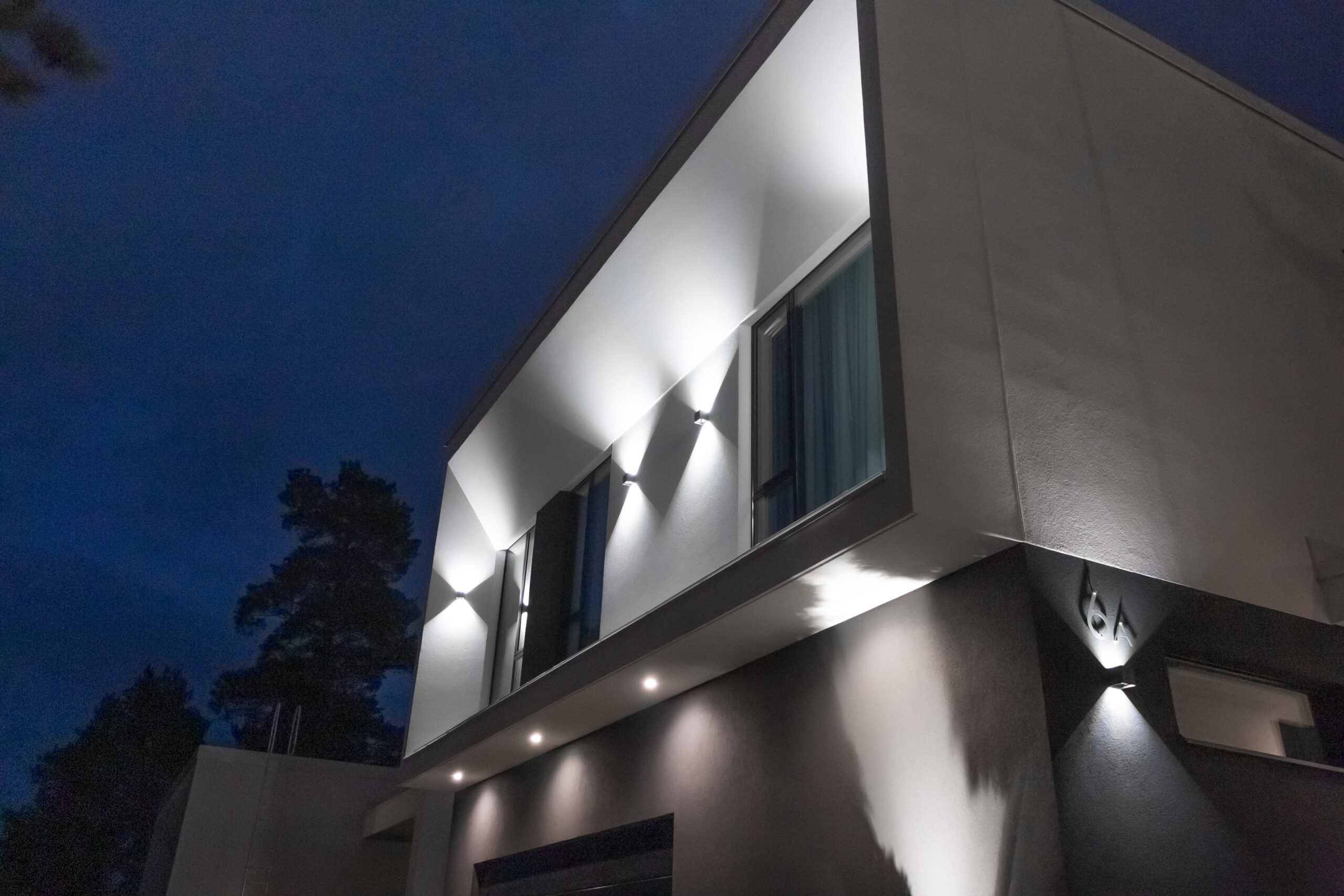 Moderni talo iltahämärässä – kulmikas arkkitehtuuri ja LedStore.fi:n laadukkaat LED-pihavalaisimet tuovat julkisivun näyttävästi esiin. - LedStore Led-pihavalaistus syntyy seinävaloista ja alaspäin osoittavista led-spoteista. Lisäksi seinävalaisin valaisee talon numeroa.