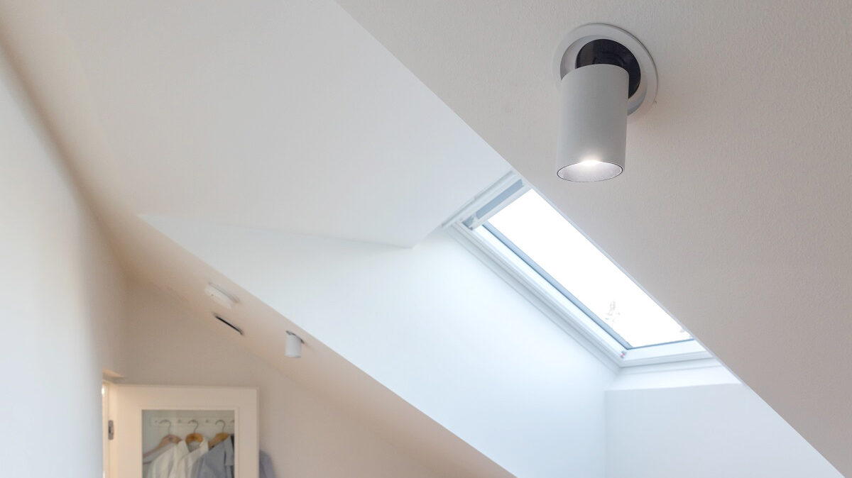 Moderni valkoinen huone, jossa LedStore.fi:n energiatehokas kattospotti ja luonnonvaloa tuova kattoikkuna. - LedStore Minimalistinen huone vinolla katolla on valaistu pyöreällä LED-kattovalaisimella sekä kattoikkunalla, valikoimaa löytyy LedStore.fi-verkkokaupasta. Kattoikkunan alla sijaitsee pieni kasvi. Vasemmalla seinällä on osittain näkyvissä ovi, josta roikkuu vaatteita. Valkoiset seinät korostavat huomioita sisustusta ja luovatin ilmapiirin.