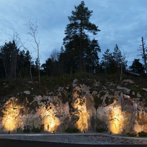 image - LedStore Yöllä kallioinen rinne kylpee lämpimässä LED-valaistuksessa, joka ruoka kivien tekstiuuria. Puut ja pensaat erottuvat rinteen huipulla vasten pilvistä, pimenevää taivasta, luoden upeaa kontrastia näkymään.