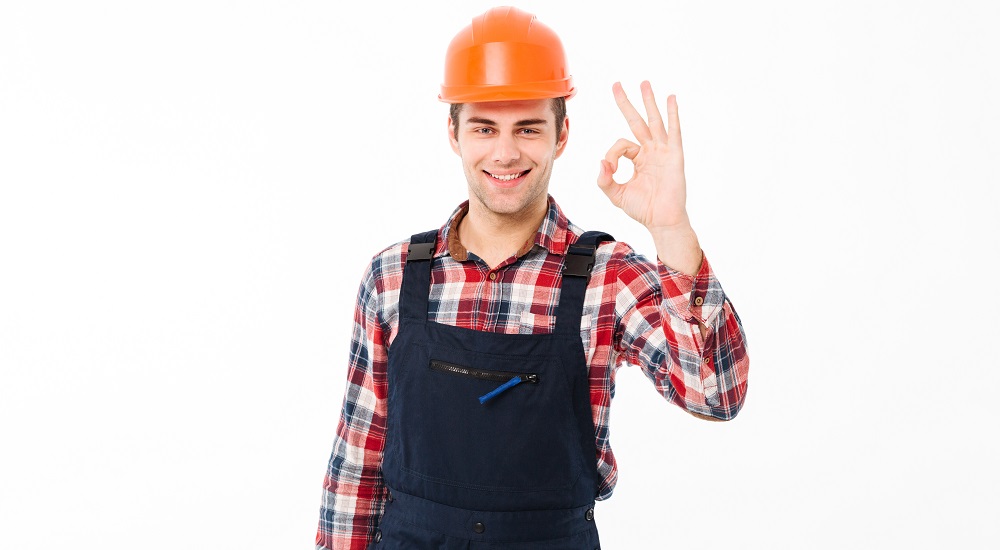 Portrait of a cheerful young male builder showing thumbs up gesture while standing and looking at camera isolated over white background.