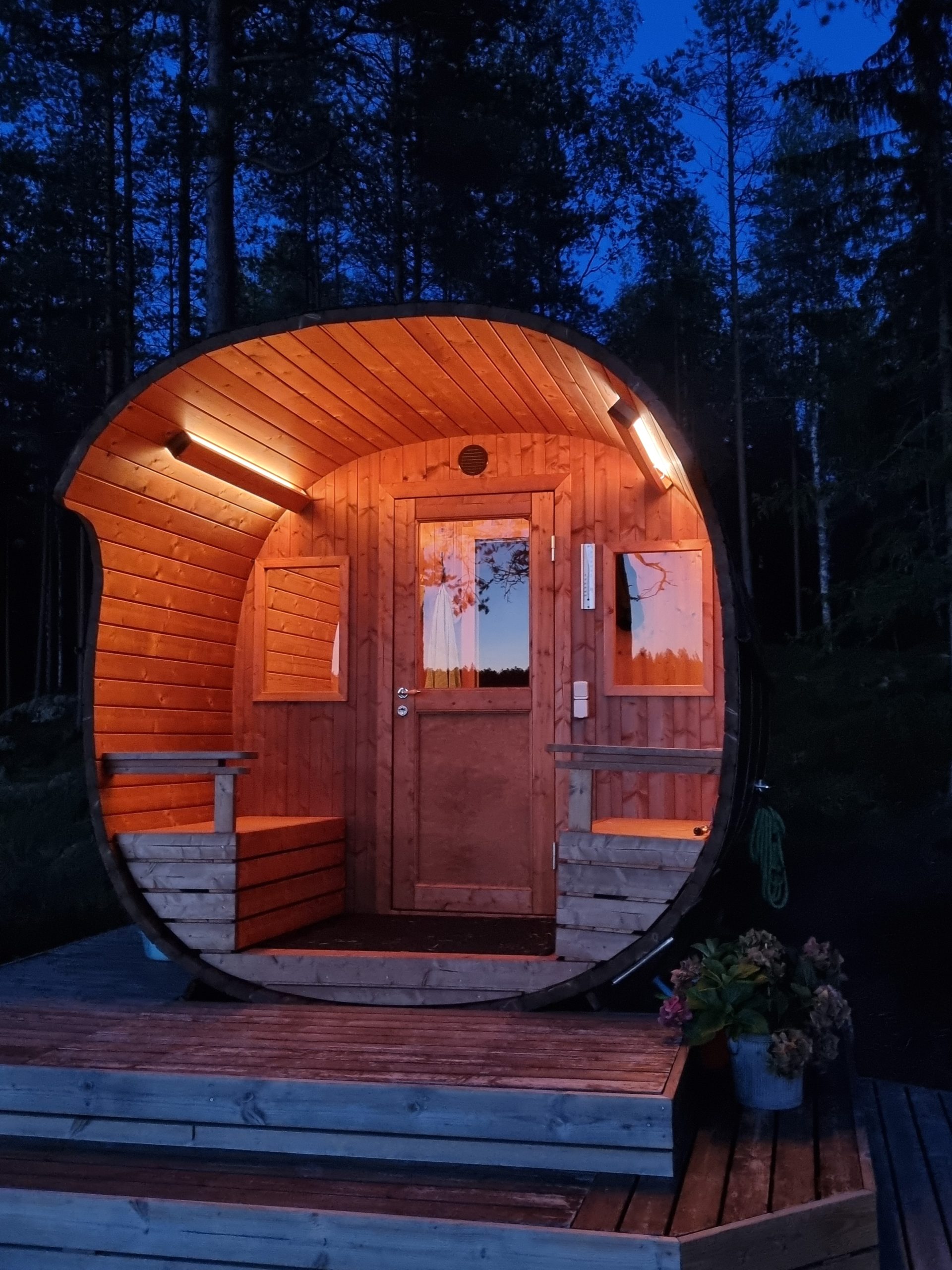Pieni kaarevamuotoinen sauna hehkuu lämpimästi illan hämärässä, kun LedStore.fi:n LED-valot korostavat tilan tunnelmaa. Se sijaitsee puisella terassilla, jota ympäröivät puut ja kukkien värit ruukussa lisää rauhallisen ympäristön viehättävyyttä.