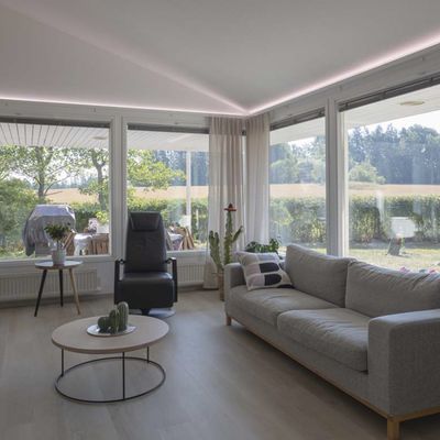 Moderni olohuone, jossa on LED-kattovalaisimet, lattiavalaisin, luonnonvaloa suurista ikkunoista, viihtyisät istuimet.