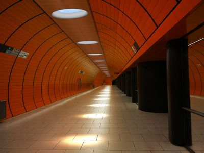 Tyhjä, hyvin valaistu oranssi metroasema, jossa on laattalattia, mustat pylväät, kaareva tunnelisuunnittelu ja seinäkyltit