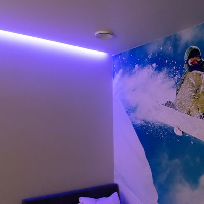 Huoneessa valkoiset seinät, violetti LED-kattovalo, seinällä hiihtäjämaalaus; näkyvä tyyny ja osa sängystä. Moderni ja viihtyisä makuuhuoneen valaistusratkaisu.