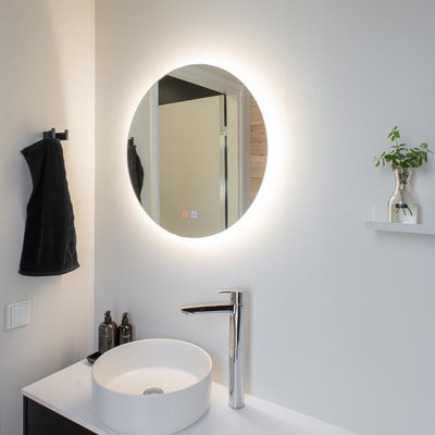 Minimalistinen kylpyhuone, jossa on LED-valaistu pyöreä peili, valkoinen astianpesuallas kromihanalla, musta pyyhe ja pieni kasvi hyllyllä.
