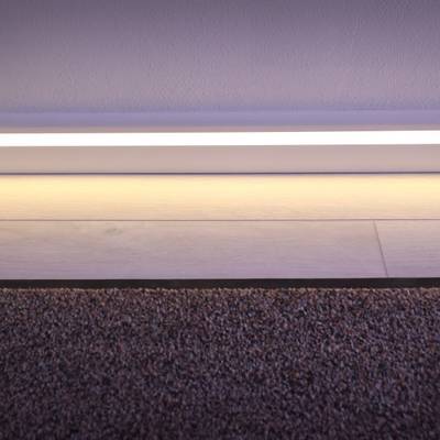 Moderni LED-nauhavalaisin valaisee siirtymän maton ja puulattian välillä korostaen valkoisia jalkalistan yksityiskohtia.