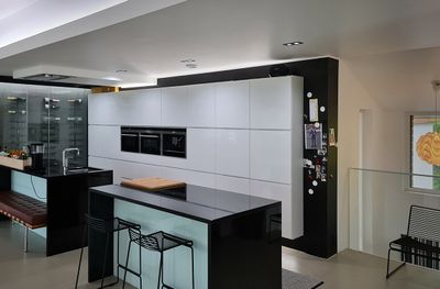 Moderni keittiö, jossa kiiltävän valkoiset kaapit, mustat työtasot, saari baarituoleilla, ruostumattomasta teräksestä valmistettu tuplauuni, jääkaappi magneeteilla oikealla LED-valaistuksen alla.