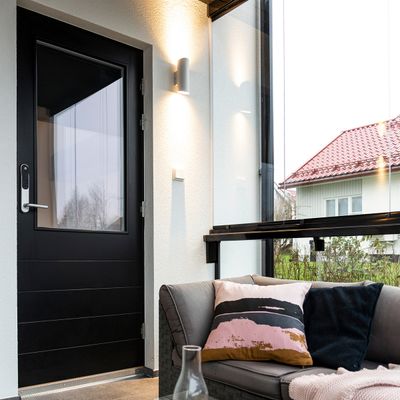 Moderni aurinkohuone, jossa on energiatehokas LED-valaistus, musta ovi, harmaa sohva värikkäillä tyynyillä, lasiseinät esittelevät taloa, jossa on eloisa punainen katto.