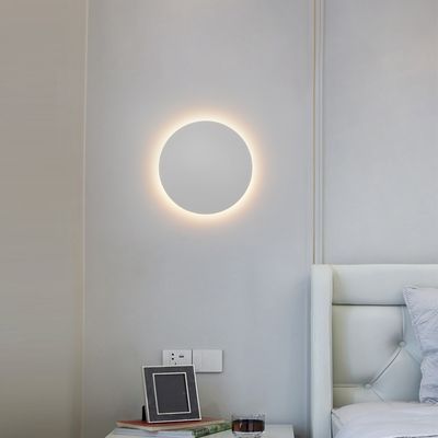 Moderni pyöreä seinävalaisin valaisee minimalistisen makuuhuoneen vaaleanharmailla seinillä, valkoisella tuftatulla sängynpäädyllä ja tyylikkäällä yöpöytäsisusuksella. LED-valaistuskauppa.
