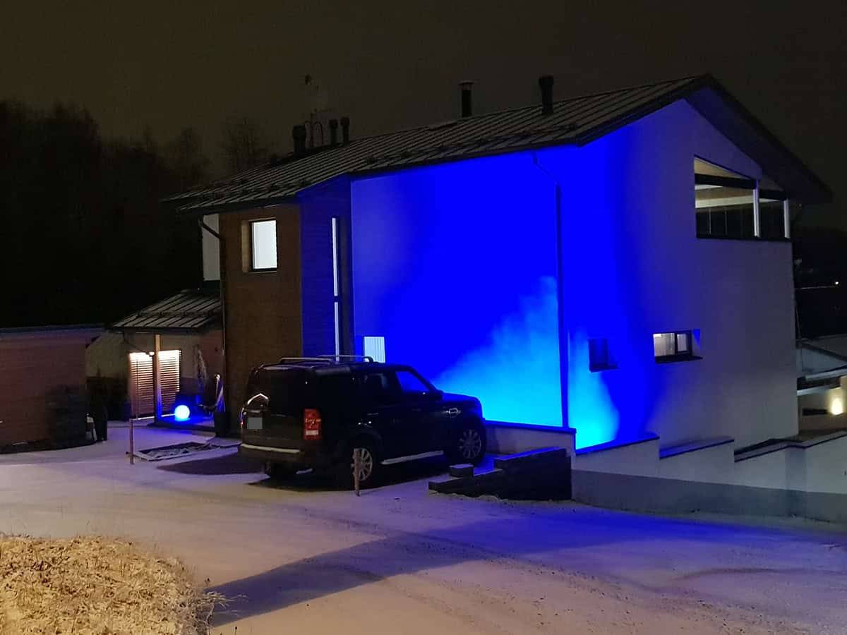 Modernin talon julkisivu valaistuna sinisillä LED-valoilla yöllä. Pihalla auto ja maassa kevyt lumipeite.