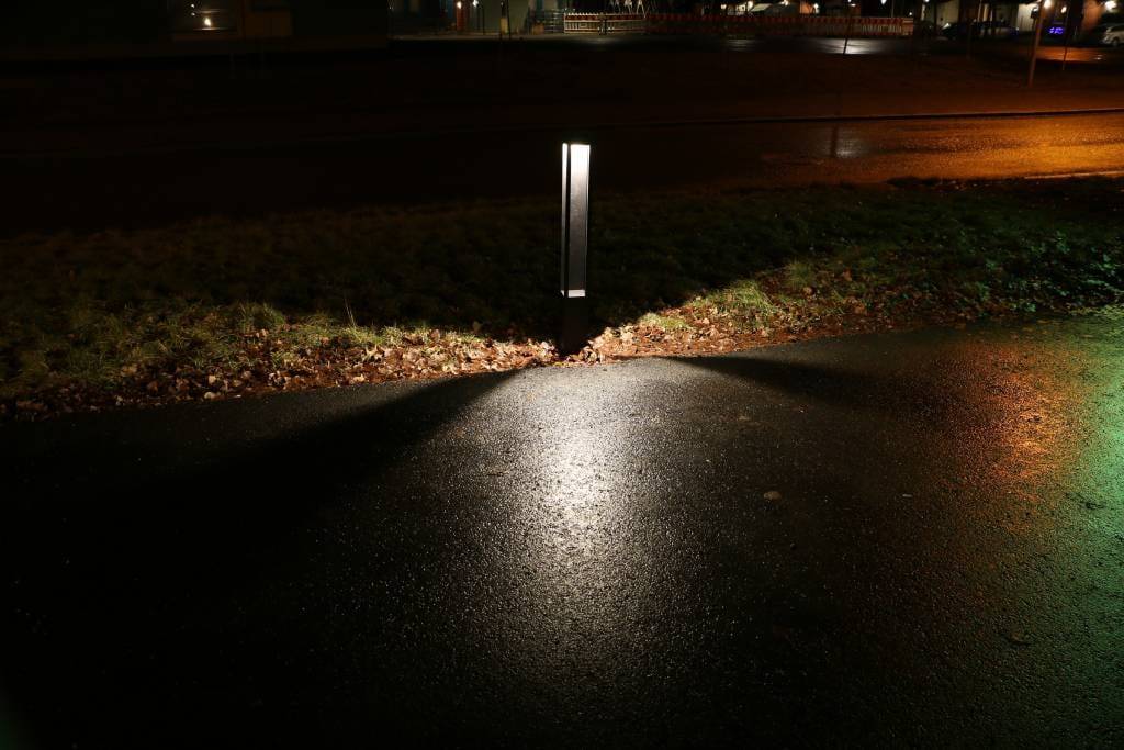 LED-polkuvalo, joka valaisee märän kadun kolmiomaisilla kuvioilla, joita reunustavat ruoho; kaupunkitausta kaukaisilla valoilla.