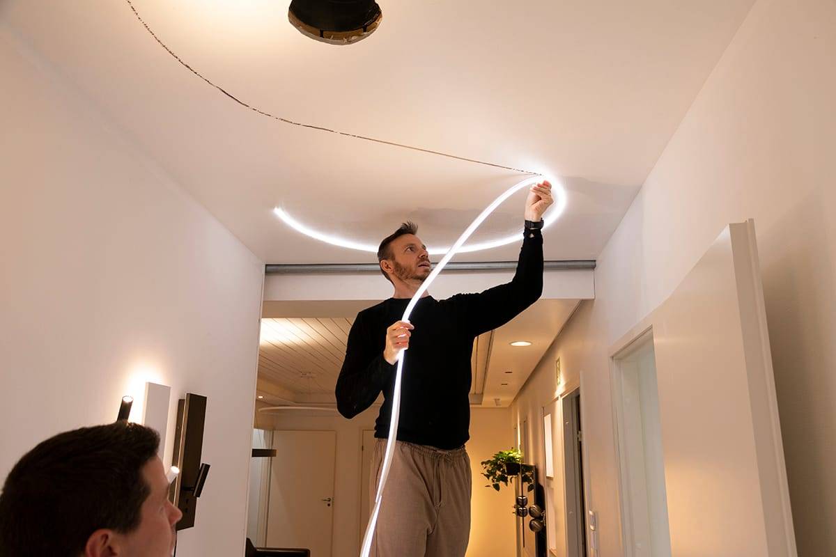 Henkilö kiinnittää joustavan LED-nauhavalaisimen moderniin kattoon, mikä parantaa huoneen valaistusta. Musta villapaita, beige housut; toinen henkilö etualalla, epätarkka.